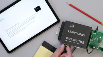 A hand wiring a North Commander unit alongside a tablet displaying a Commander Tutorial document, a notepad, and a screwdriver on a desk.