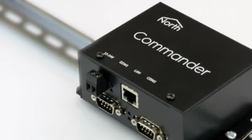 Black North Commander building controller unit mounted on a metal DIN rail, showing power input, serial, and LAN connection ports.
